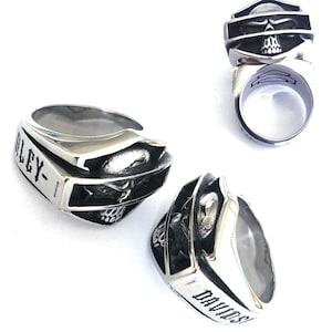 Peut inclure: Une bague argentée avec un motif de crâne et les mots "HARLEY-DAVIDSON" gravés sur les côtés. La bague a un dessus en forme de bouclier avec un visage de crâne. La bague est montrée sous plusieurs angles.