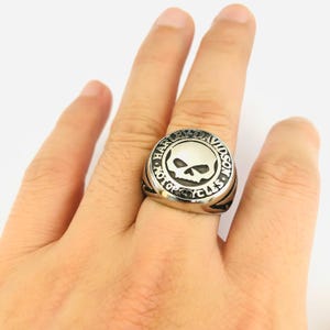 May include: A silver ring with a skull emblem at the centre, encircled by the words "HARLEY-DAVIDSON MOTORCYCLES". The ring's design includes a wide band with grooved detailing. The ring is a motorcycle-themed accessory.