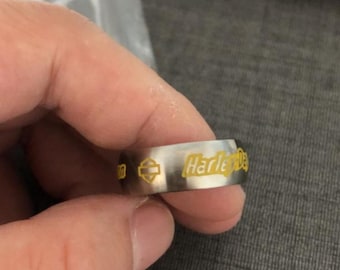May include: A silver-toned metal band ring featuring a brushed finish. The exterior surface is engraved with the yellow text Harley-Davidson and a yellow bar and shield logo. The ring has a smooth, flat profile and a consistent width throughout.