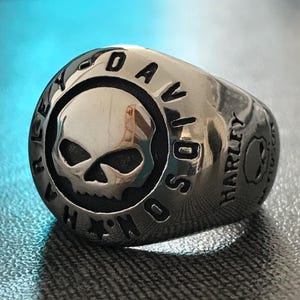 Op de afbeelding: Een zilverkleurige ring met een schedelontwerp in het midden. De ring is gegraveerd met de woorden "HARLEY DAVIDSON" rond de schedel en aan de zijkant. De schedel heeft gedetailleerde kenmerken en de ring heeft een gepolijste afwerking.