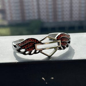 Puede incluir: Brazalete plateado con un emblema central y alas adornadas con piedras rojas. El brazalete tiene un acabado pulido. El diseño es audaz y llamativo, ideal para añadir un toque distintivo.