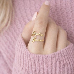 Può includere: Un anello dorato con i nomi "Sophia" e "Gavin" scritti in corsivo. L'anello è indossato su un dito, con i nomi disposti verticalmente. Lo sfondo è di colore rosa tenue.