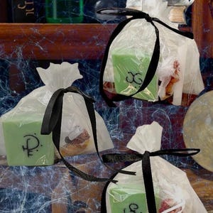Puede incluir: Jabones verdes en forma de cubo con símbolos negros están empaquetados en bolsas de celofán transparente y atados con cintas negras. El fondo incluye una estantería con botellas y libros.