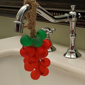 May include: A decorative cluster of translucent red and green spherical objects resembling grapes, suspended from a chrome faucet by a thick, natural-colored jute rope. The fruit-shaped items have a matte, frosted texture and are arranged in a bunch.