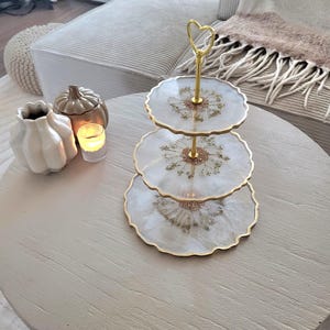Könnte beinhalten: Ein dreistufiges Serviertablett mit goldfarbenem Stiel und herzförmigem Griff. Jede Ebene ist ein Kreis aus klarem Harz mit gewelltem Rand, eingebetteten Trockenblumen und goldenem Rand. Das Tablett steht auf einem hellen runden Tisch.