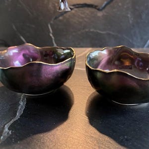 Op de afbeelding: Twee kleine decoratieve kommen met een donkere, iriserende afwerking, met tinten paars en groen. De randen zijn afgewerkt met goud. Deze unieke kommen zijn geschikt voor het serveren van snacks of als decoratieve items.