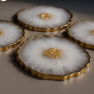 Op de afbeelding: Vier decoratieve onderzetters met een wit, gemarmerd ontwerp en goudkleurige randen. Elke onderzetter heeft een centrale cluster van goudkleurige korrels, wat een elegante uitstraling creëert. De onderzetters zijn op een neutraal oppervlak geplaatst.