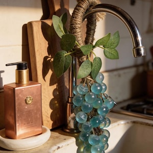 Op de afbeelding: Een koperkleurige zeepdispenser met een gouden pomp staat op een wit keramisch schaaltje. Een decoratieve tros blauwe glazen druiven hangt aan de kraan, versierd met groene bladeren. Een houten snijplank staat op de achtergrond.