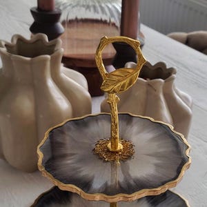 Op de afbeelding: Een serveerschaal met twee niveaus met een goudkleurige steel en een blad-vormige handgreep. De schalen hebben een zwart-wit marmerpatroon met gouden rand. Decoratieve vazen en kaarsen staan op de achtergrond.