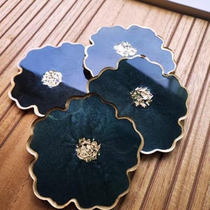Puede incluir: Cuatro posavasos de resina con diseño floral. Cada posavasos es de color azul verdoso oscuro con un borde dorado y un centro dorado. Los posavasos están dispuestos sobre una superficie de madera.