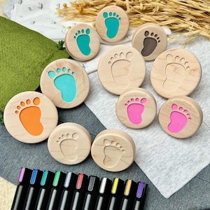 May include: A collection of round wooden stamps featuring engraved baby footprint designs. Some stamps have colorful inserts in orange, teal, and pink, while others are plain wood. A row of black markers with colored caps sits below.