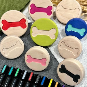 Puede incluir: Una colección de discos de madera redondos, cada uno con una forma de hueso tallada. Algunos recortes de hueso están rellenos con colores vibrantes, incluyendo rojo, rosa, verde lima, azul y negro, mientras que otros permanecen en madera natural.