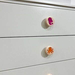 May include: A white dresser featuring three round wooden drawer knobs. Each knob displays a colorful cutout design: a magenta mermaid, an orange octopus, and a dark blue seahorse, all set against the smooth white surface of the furniture.