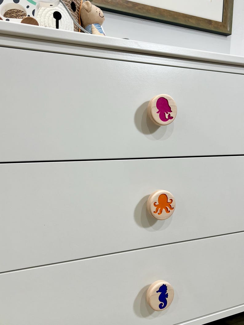 May include: A white dresser featuring three round wooden drawer knobs. Each knob displays a colorful cutout design: a magenta mermaid, an orange octopus, and a dark blue seahorse, all set against the smooth white surface of the furniture.