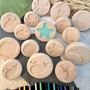 May include: A collection of round wooden discs featuring engraved marine life designs including an octopus, seahorse, whale, dolphin, and seashell. One disc features a central teal starfish cutout. A set of black markers is positioned below.