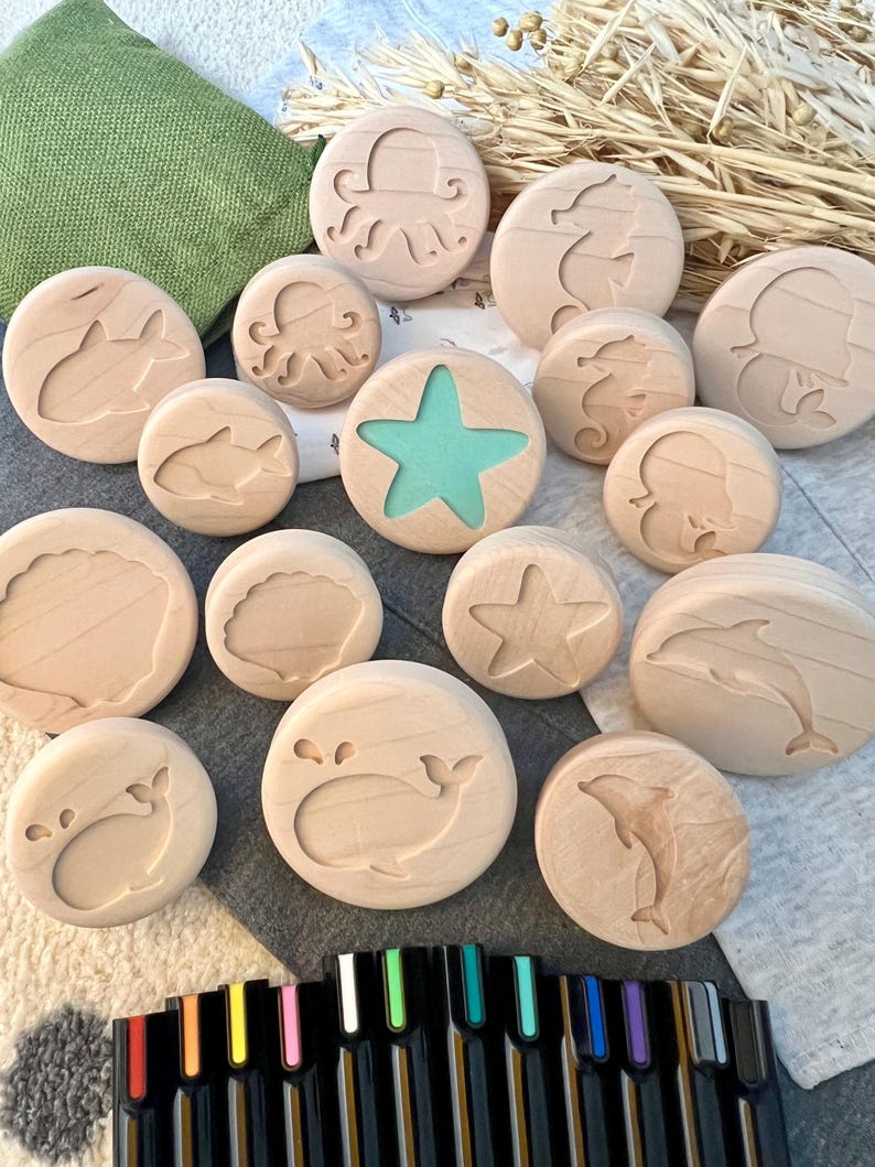 May include: A collection of round wooden discs featuring engraved marine life designs including an octopus, seahorse, whale, dolphin, and seashell. One disc features a central teal starfish cutout. A set of black markers is positioned below.