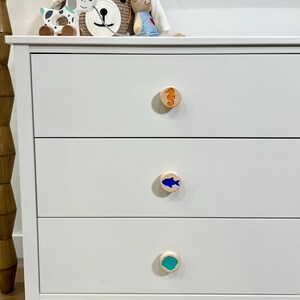 May include: A white three-drawer dresser featuring round wooden knobs. The top knob displays an orange seahorse, the middle knob features a blue fish, and the bottom knob shows a teal shell shape. The dresser has a smooth, matte finish.