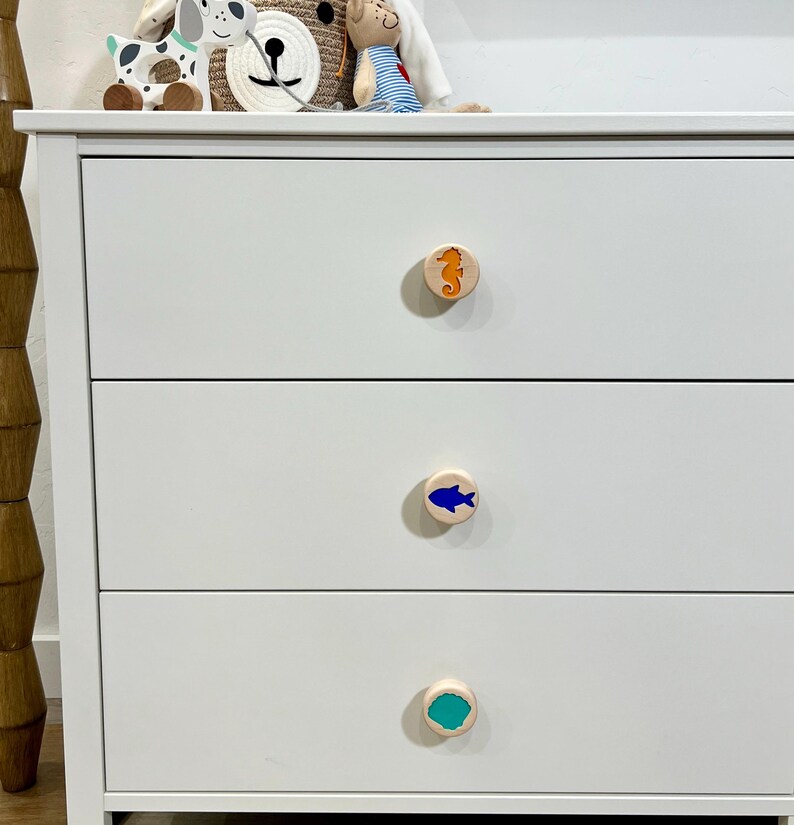 May include: A white three-drawer dresser featuring round wooden knobs. The top knob displays an orange seahorse, the middle knob features a blue fish, and the bottom knob shows a teal shell shape. The dresser has a smooth, matte finish.