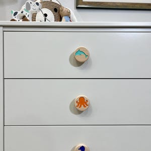 May include: A white three-drawer dresser featuring three circular wooden knobs. Each knob displays a different colored sea creature inlay: a teal dolphin on the top, an orange octopus in the middle, and a dark blue seahorse on the bottom drawer.