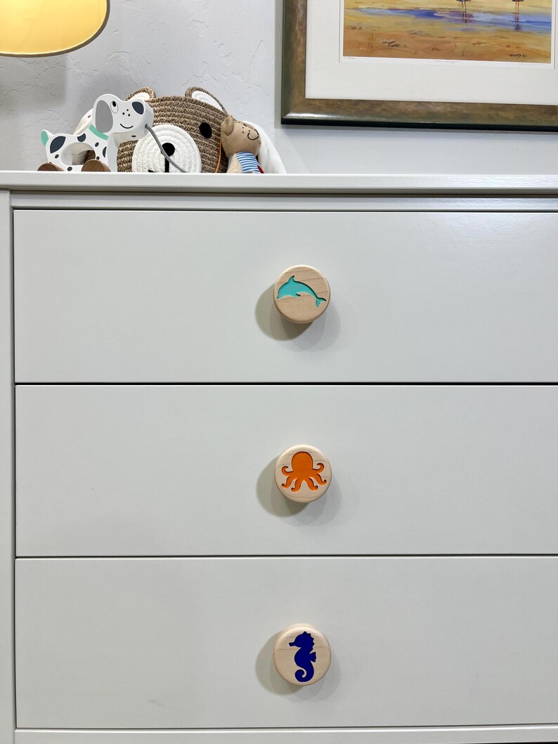 May include: A white three-drawer dresser featuring three circular wooden knobs. Each knob displays a different colored sea creature inlay: a teal dolphin on the top, an orange octopus in the middle, and a dark blue seahorse on the bottom drawer.