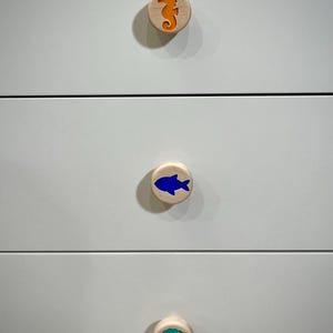 May include: Three circular light-colored wooden drawer knobs attached to a white dresser. The top knob features an orange seahorse inlay, the middle knob has a blue fish inlay, and the bottom knob displays a teal seashell inlay.