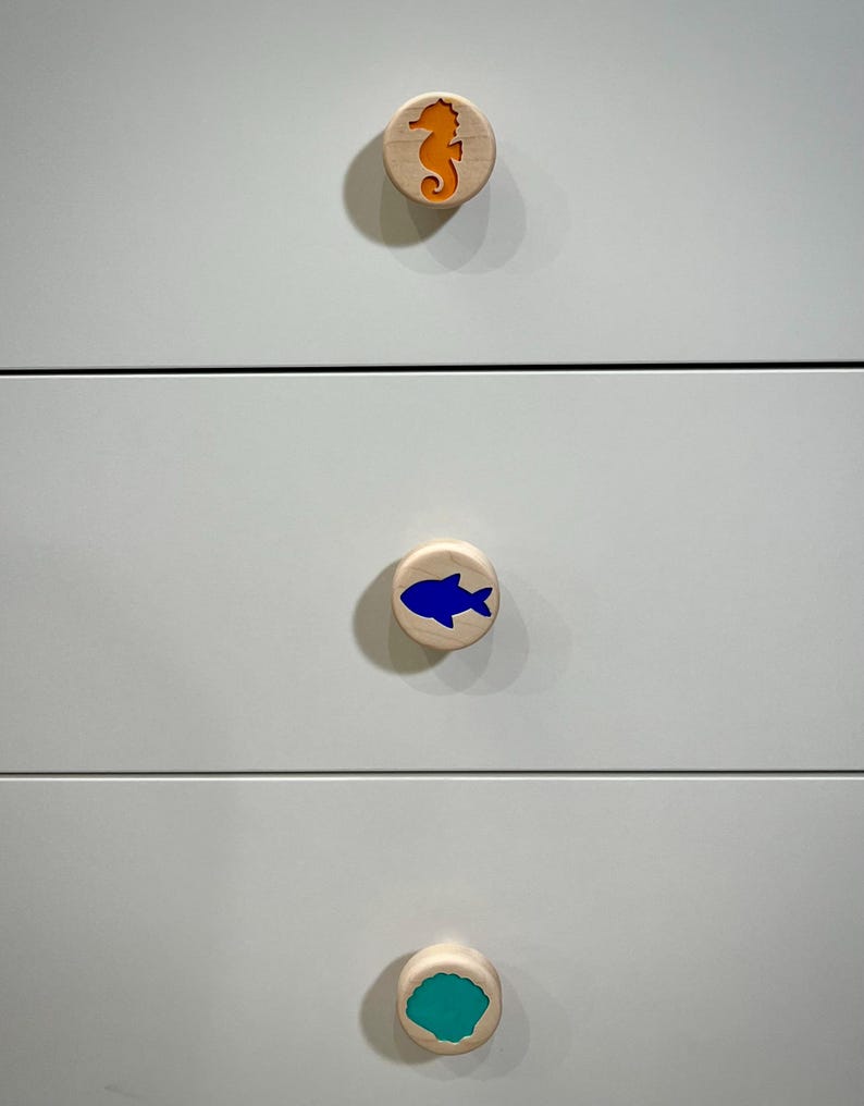 May include: Three circular light-colored wooden drawer knobs attached to a white dresser. The top knob features an orange seahorse inlay, the middle knob has a blue fish inlay, and the bottom knob displays a teal seashell inlay.