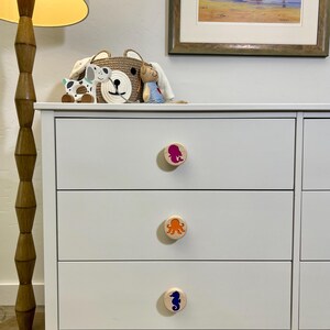 May include: A white three-drawer dresser featuring circular wooden knobs. Each knob is decorated with a colorful sea creature graphic: a magenta mermaid on the top, an orange octopus in the middle, and a blue seahorse on the bottom drawer.