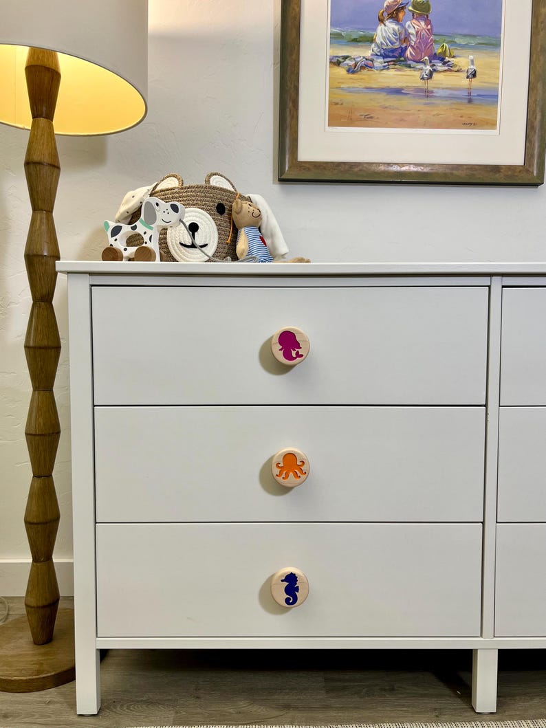 May include: A white three-drawer dresser featuring circular wooden knobs. Each knob is decorated with a colorful sea creature graphic: a magenta mermaid on the top, an orange octopus in the middle, and a blue seahorse on the bottom drawer.