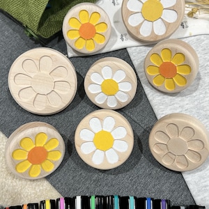 May include: A collection of round wooden stamps featuring flower designs, some with yellow and white paint and others left plain. The set includes several circular stamps with carved daisy patterns, alongside a row of black markers with colored caps.