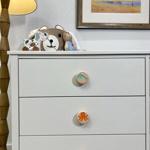 May include: A white three-drawer dresser featuring circular wooden knobs. Each knob displays a different colored sea creature graphic: a light blue dolphin on the top, an orange octopus in the middle, and a dark blue seahorse on the bottom.