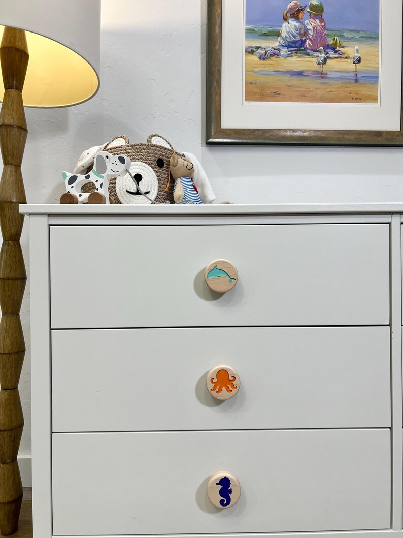 May include: A white three-drawer dresser featuring circular wooden knobs. Each knob displays a different colored sea creature graphic: a light blue dolphin on the top, an orange octopus in the middle, and a dark blue seahorse on the bottom.