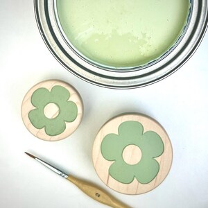 May include: A metal paint can filled with light green paint, two round wooden discs featuring a light green flower inlay, and a thin wooden paintbrush with the number 2 and the text YFLLN printed on the handle, all arranged on a white surface.