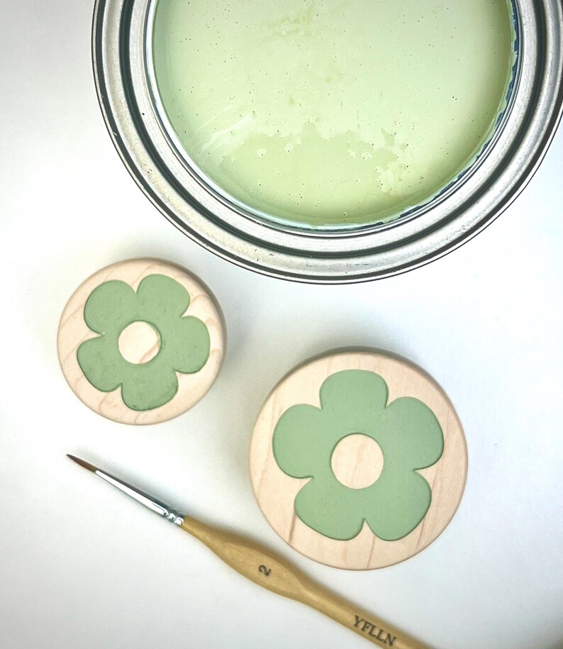 May include: A metal paint can filled with light green paint, two round wooden discs featuring a light green flower inlay, and a thin wooden paintbrush with the number 2 and the text YFLLN printed on the handle, all arranged on a white surface.