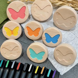 May include: A collection of nine circular wooden stamps, each featuring a butterfly cutout. Some stamps have colored butterfly inserts in pink, yellow, orange, blue, and teal, while others are plain wood. A row of black markers is positioned below.