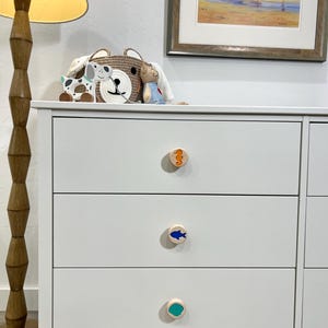 May include: A white dresser with three drawers featuring round wooden knobs. Each knob displays a different colored icon: an orange seahorse on the top, a blue fish in the middle, and a solid teal circle on the bottom drawer knob.