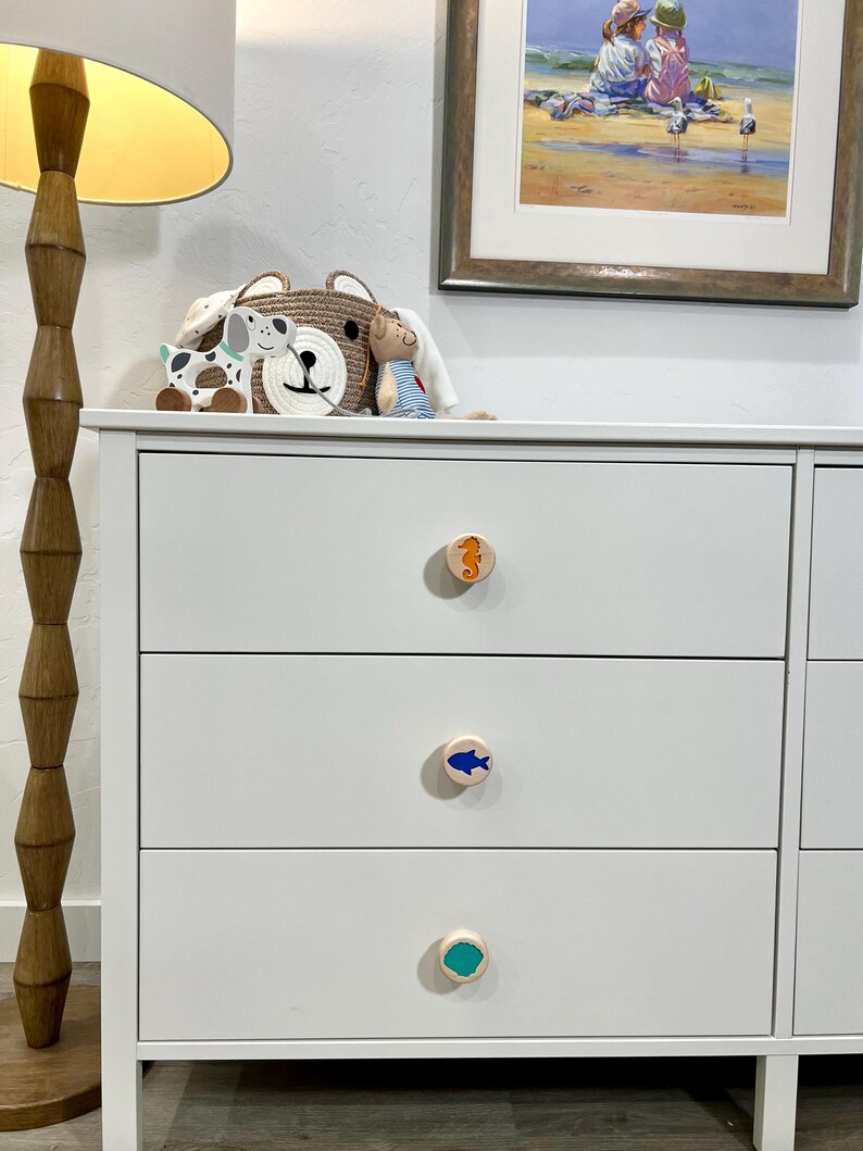 May include: A white dresser with three drawers featuring round wooden knobs. Each knob displays a different colored icon: an orange seahorse on the top, a blue fish in the middle, and a solid teal circle on the bottom drawer knob.