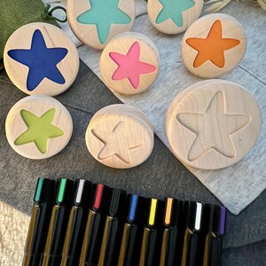 May include: A collection of round wooden discs featuring star-shaped cutouts, some filled with blue, teal, pink, orange, or lime green resin, alongside others left as plain wood. Below the discs are several black markers with colored caps.