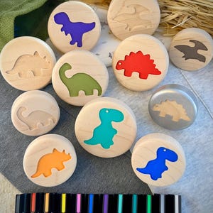 May include: A collection of round wooden discs featuring various dinosaur silhouettes. Some discs have colorful inlays in purple, green, red, teal, orange, and blue, while others show natural wood or metallic finishes. A row of black markers is positioned below.