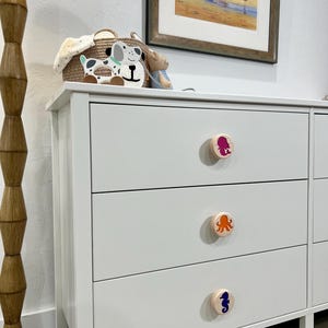 May include: A white three-drawer dresser featuring round wooden knobs. Each knob displays a colorful graphic: a pink mermaid on the top, an orange octopus in the middle, and a dark blue seahorse on the bottom drawer. The dresser has a smooth finish.
