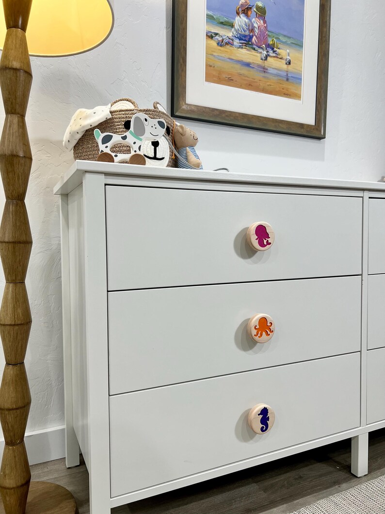 May include: A white three-drawer dresser featuring round wooden knobs. Each knob displays a colorful graphic: a pink mermaid on the top, an orange octopus in the middle, and a dark blue seahorse on the bottom drawer. The dresser has a smooth finish.