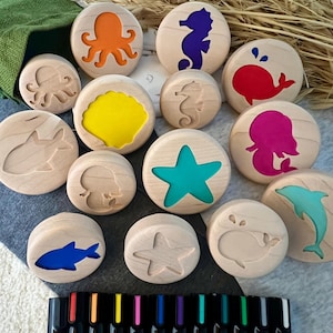 May include: A collection of round wooden stamps featuring various sea creature silhouettes including an octopus, seahorse, whale, shark, starfish, and mermaid. Some stamps have colorful inlays while others are plain wood. A set of black markers is included.