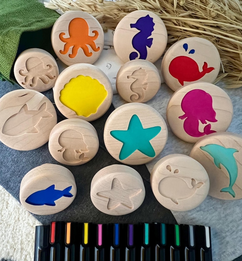 May include: A collection of round wooden stamps featuring various sea creature silhouettes including an octopus, seahorse, whale, shark, starfish, and mermaid. Some stamps have colorful inlays while others are plain wood. A set of black markers is included.