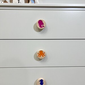 May include: A white dresser with three drawers, each featuring a round, light-colored wooden knob. The top knob displays a magenta mermaid silhouette, the middle knob shows an orange octopus, and the bottom knob features a blue seahorse.