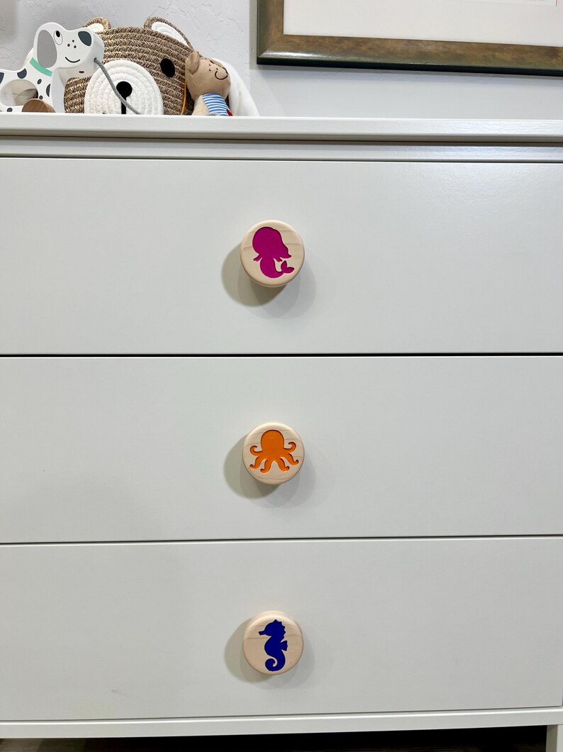 May include: A white dresser with three drawers, each featuring a round, light-colored wooden knob. The top knob displays a magenta mermaid silhouette, the middle knob shows an orange octopus, and the bottom knob features a blue seahorse.