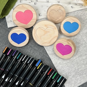 May include: A collection of six circular wooden stamps featuring heart-shaped centers, with three stamps displaying pink, blue, and purple ink, alongside a set of black Sharpie markers with various colored caps arranged in a row.