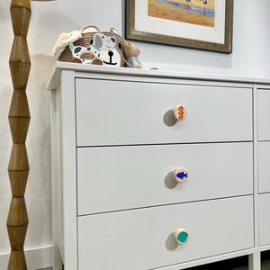 May include: A white dresser featuring three drawers, each equipped with a round wooden knob. The top knob displays an orange seahorse, the middle knob features a blue fish, and the bottom knob shows a teal circular design.