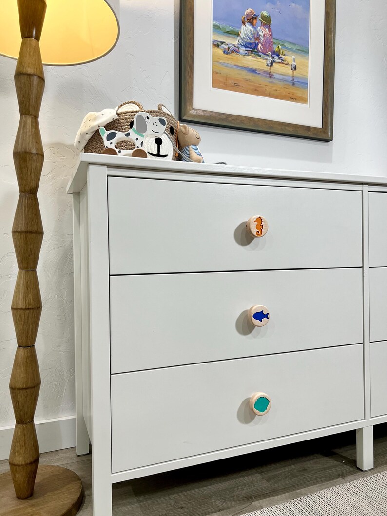 May include: A white dresser featuring three drawers, each equipped with a round wooden knob. The top knob displays an orange seahorse, the middle knob features a blue fish, and the bottom knob shows a teal circular design.