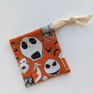 Puede incluir: Una bolsa de tela naranja con un patrón repetido de calaveras blancas, fantasmas, murciélagos y otras imágenes temáticas de Halloween. Tiene un cierre de cordón de color crema.