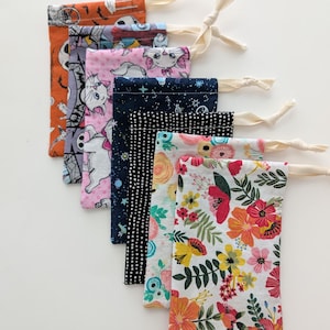 May include: A collection of seven fabric drawstring bags with various patterns. The patterns include Halloween-themed designs, cat illustrations, a starry night, polka dots, and floral prints. Each bag has a cream-colored drawstring.