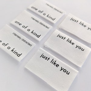 K&ouml;nnte beinhalten: Sechs wei&szlig;e Stoff-Etiketten mit schwarzem Text. Die Etiketten lauten "one of a kind" und "just like you".
